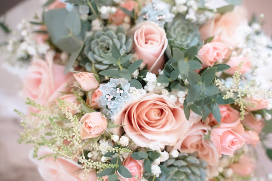 A close-up view of a floral arrangement featuring soft peach roses, delicate pink spray roses, and clusters of white baby's breath. Interspersed with green eucalyptus leaves and silvery succulents, th