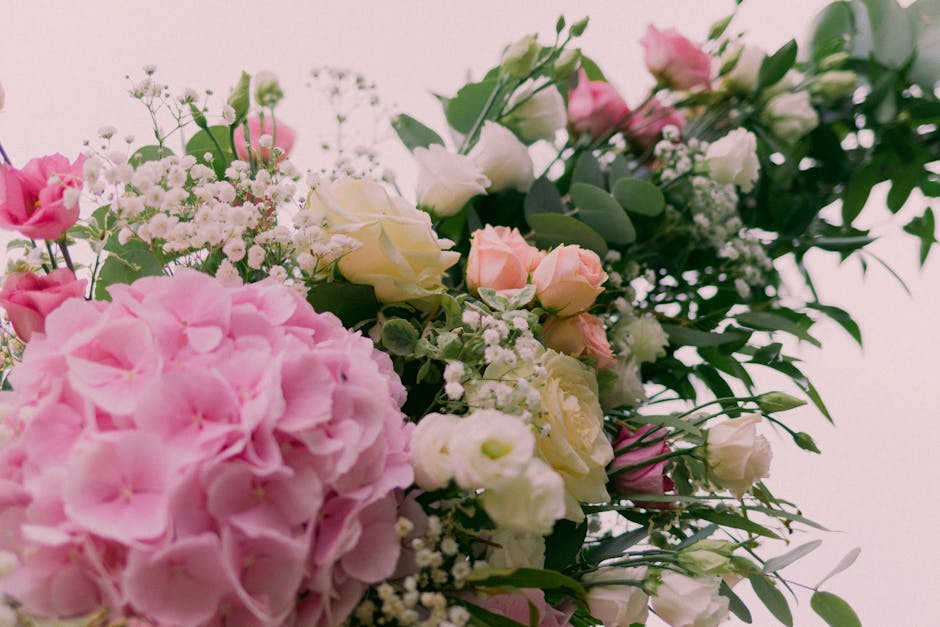 A close-up view of a floral arrangement featuring large pink hydrangea blossoms, white and soft pink roses, and delicate sprigs of baby's breath, all nestled amid lush green foliage. The flowers are a
