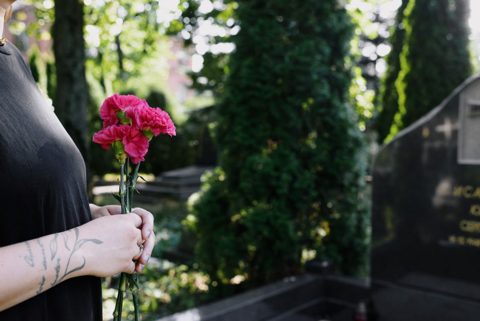 A person dressed in dark clothing holds a small bouquet of vibrant pink carnations with green stems wrapped in clear plastic. The background features a green, leafy cemetery scene with sunlight filter