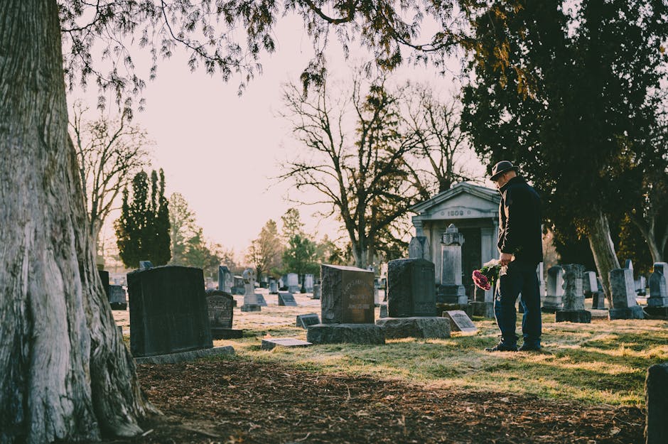 A man dressed in dark formal clothing and a hat, standing in a cemetery at sunset while holding a floral arrangement of pink and red flowers. The cemetery features numerous upright and leaning tombsto